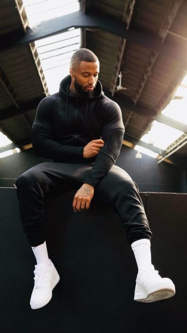 A muscular man in a black hoodie and joggers sitting on a ledge in an industrial space.